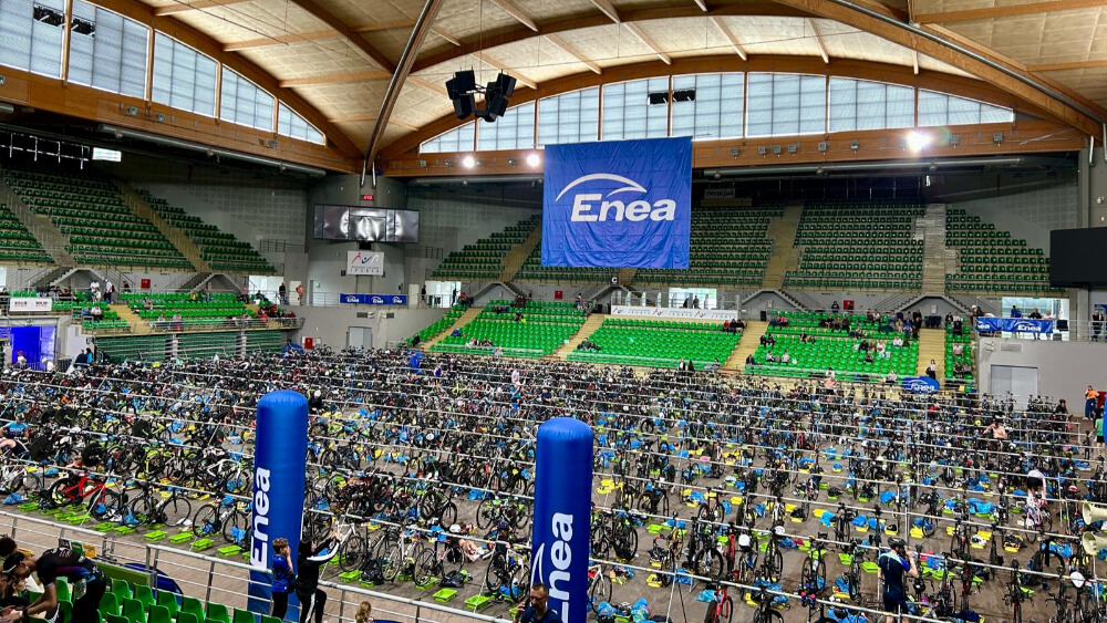 Hala sportowa z zielonymi trybunami i setkami rowerów w strefie zmian triathlonu, z dużym banerem Enea zawieszonym nad areną
